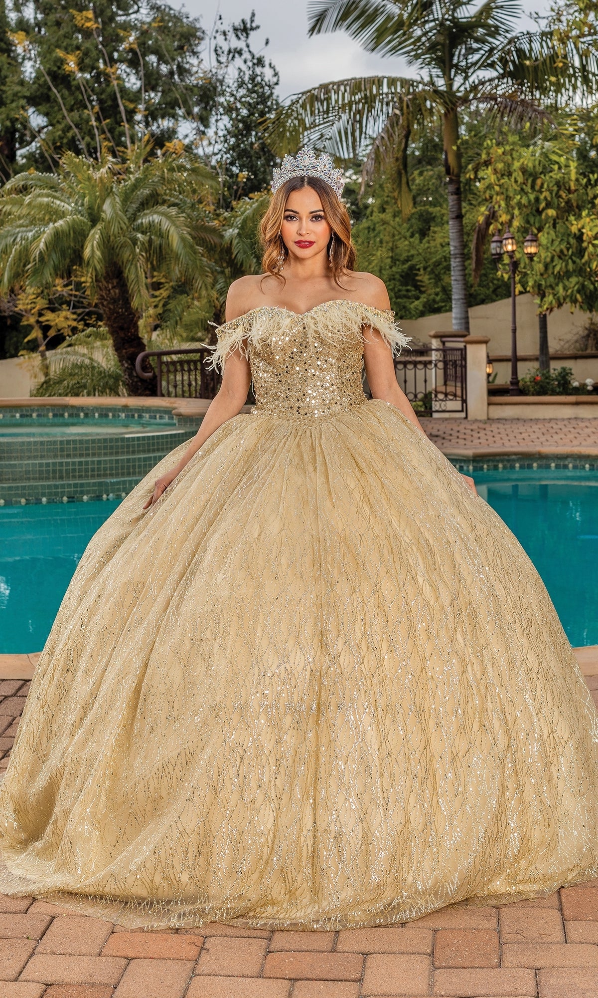 A woman wearing the Quinceanera Dress 1829 by Dancing Queen and a tiara stands in front of a swimming pool, surrounded by palm trees and lush greenery.